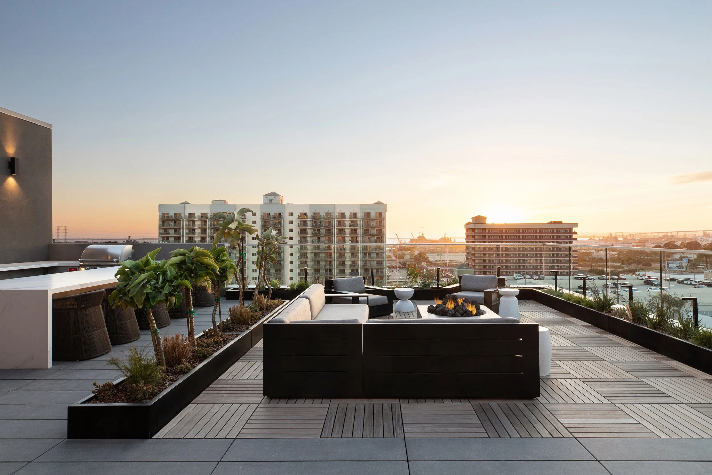 Multifamily rooftop amenity terrace with outdoor lounge seating, fire pit, landscaped planters, and views of San Diego at sunset.