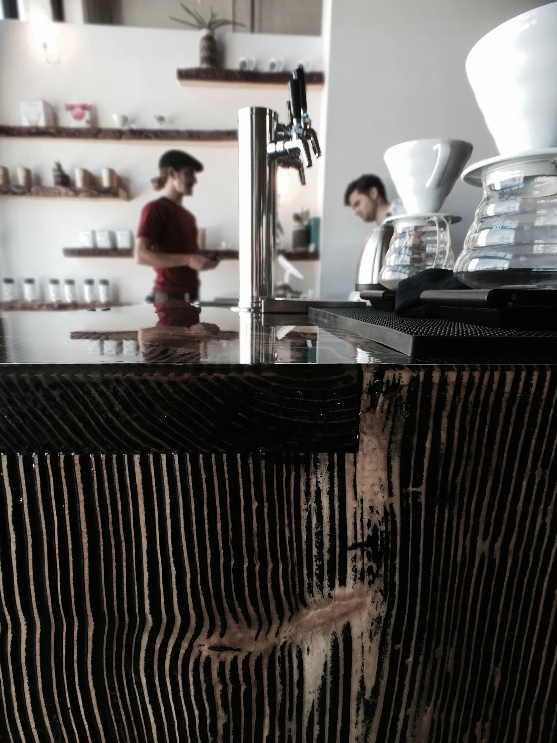 Baristas at a charred timber coffee counter with brewing equipment and cup shelving