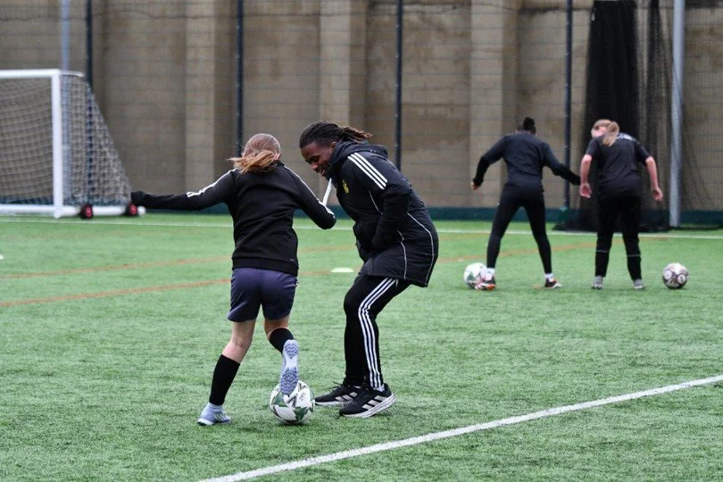 A coach in a black adidas tracksuit working with a young player on the ball during an outdoor training session
