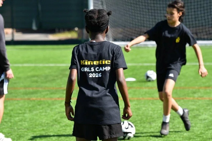Girls playing football on a grass pitch wearing Kinetic Girls Camp shirts