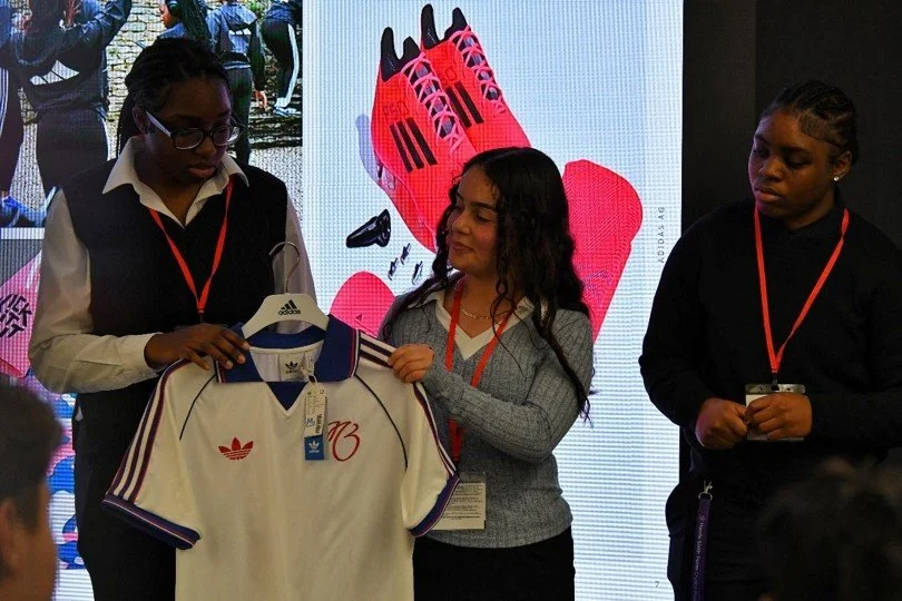 Students holding an adidas shirt in front of a display screen during a workplace visit