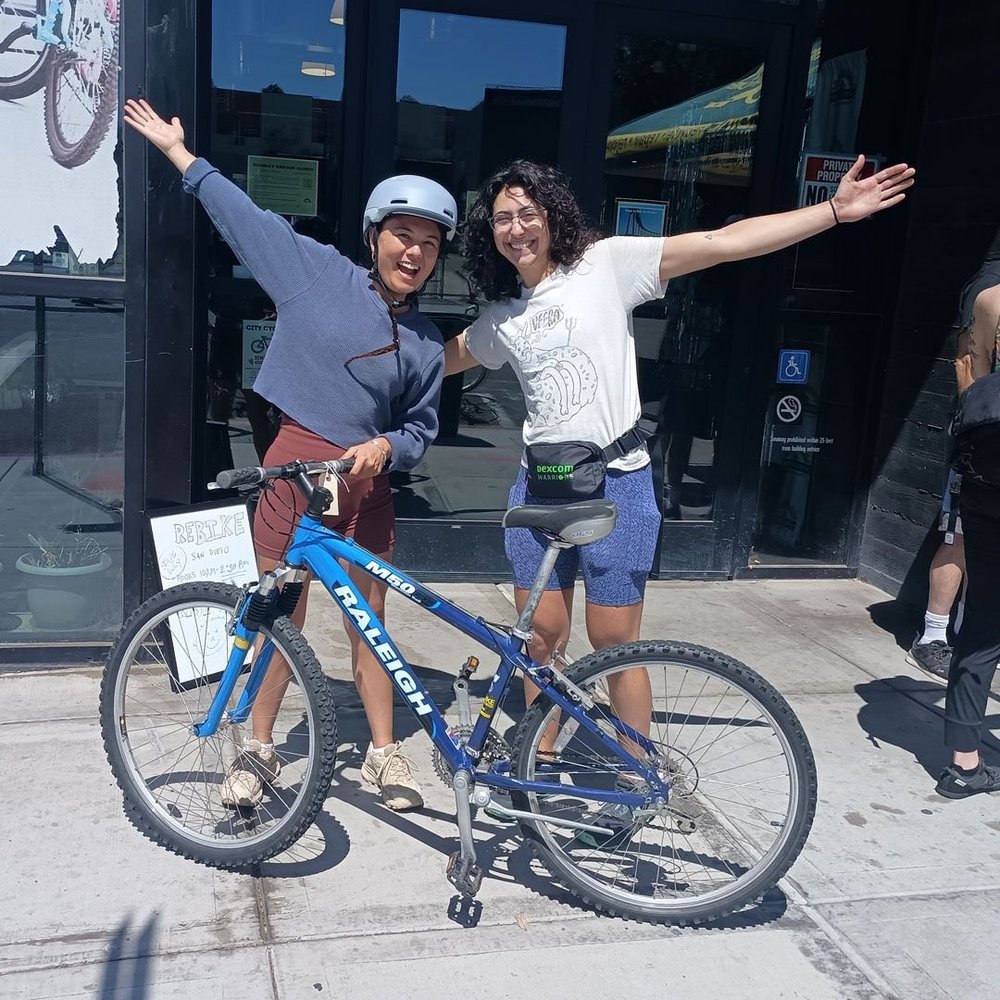 Two riders throw their arms up beside a newly refurbished blue bike