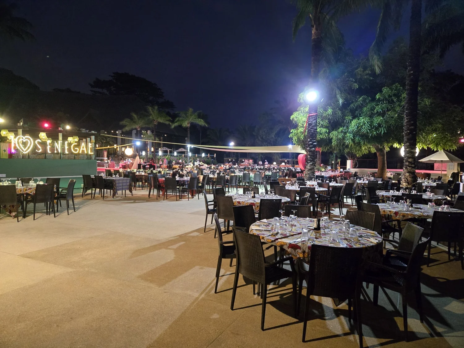 Les tables du dîner, nappes colorées, au bord de la piscine le soir, sous les palmiers et l’enseigne lumineuse « SENEGAL », au Club Med Cap Skirring (Sénégal)
