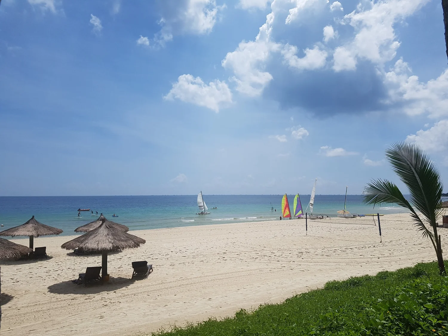 La plage de sable blanc, ses parasols de paille, un filet de volley et des catamarans aux voiles colorées au bord de l’eau, au Club Med Bintan Island (Indonésie)