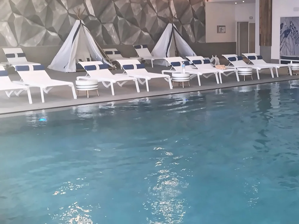The indoor pool: white loungers with blue-striped cushions and two white tepees against a faceted grey wall, beside clear blue-green water