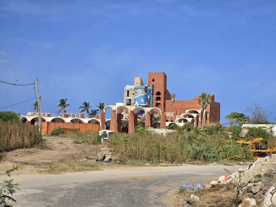 Les arches et les tours de brique de l’ancien Club Med Les Almadies à Dakar, au Sénégal, avec un panneau de mosaïque bleue, envahies par les herbes hautes, avec un conteneur et un engin de chantier jaune au premier plan, en 2025