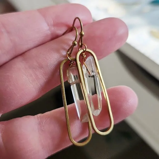 Hand-held close-up of wire-wrapped clear quartz earrings in a brass oval setting.