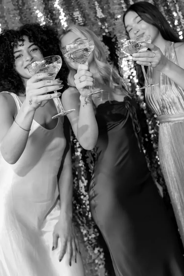 Black and white photo of three guests raising coupe glasses in front of a sequin backdrop