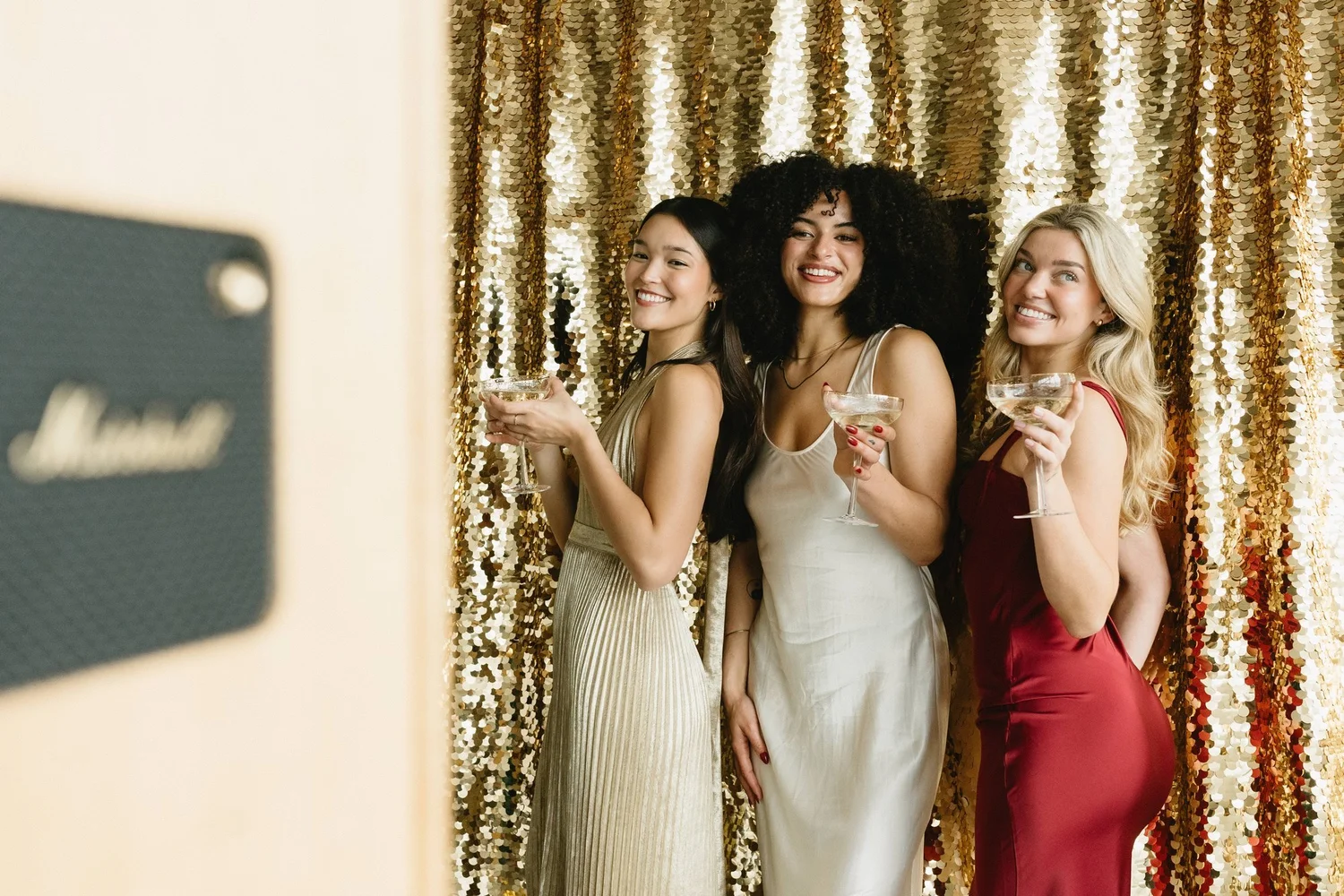 Three guests with champagne at a holiday party photo booth in front of a gold sequin backdrop