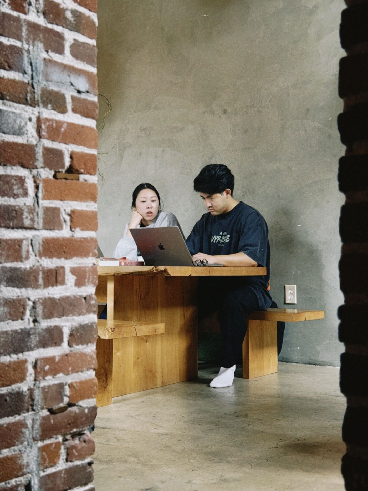 Two people working together on a laptop at a wood table in a brick-and-concrete studio