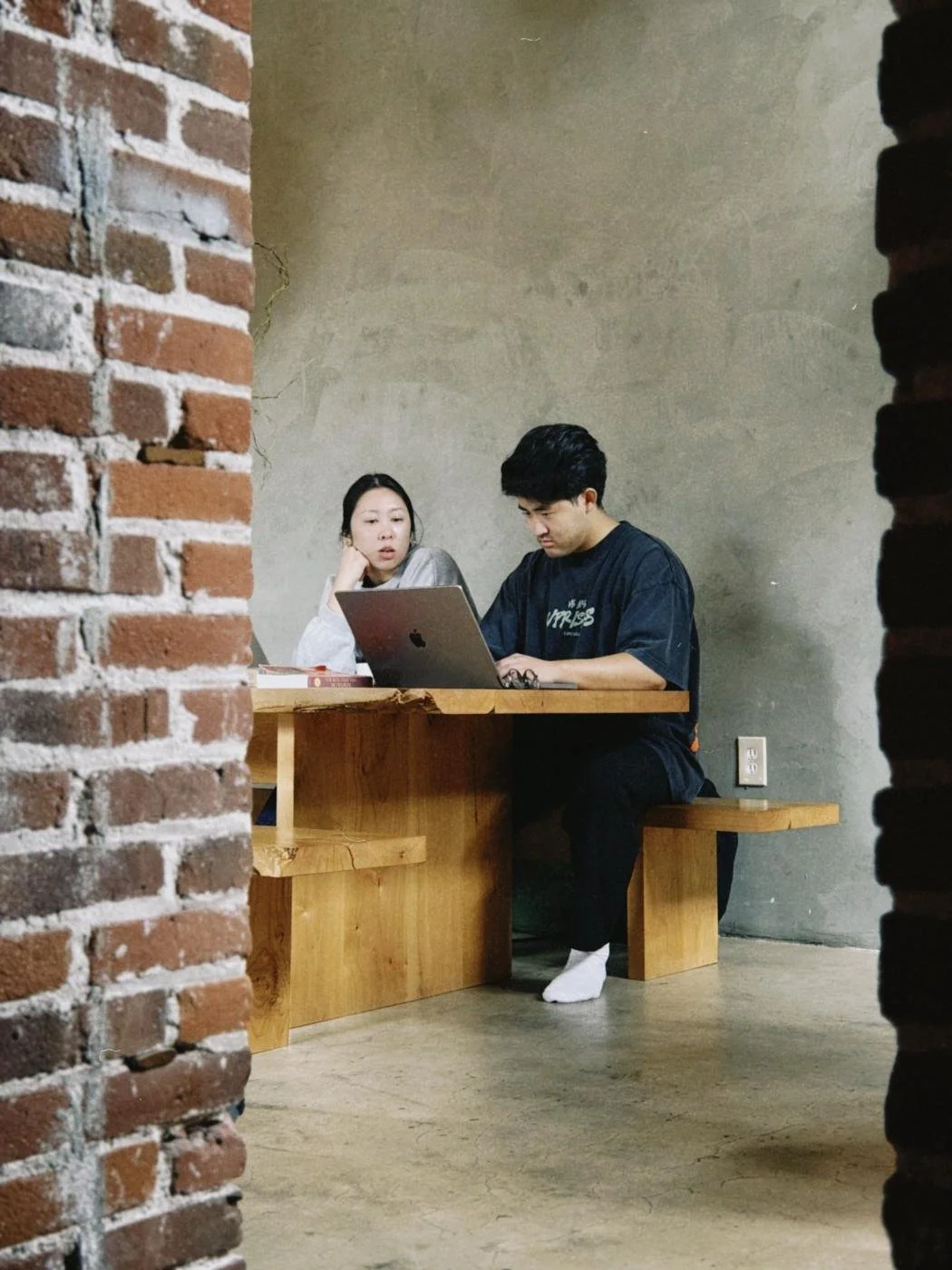 Two people working together at a wood table in a brick-and-concrete studio