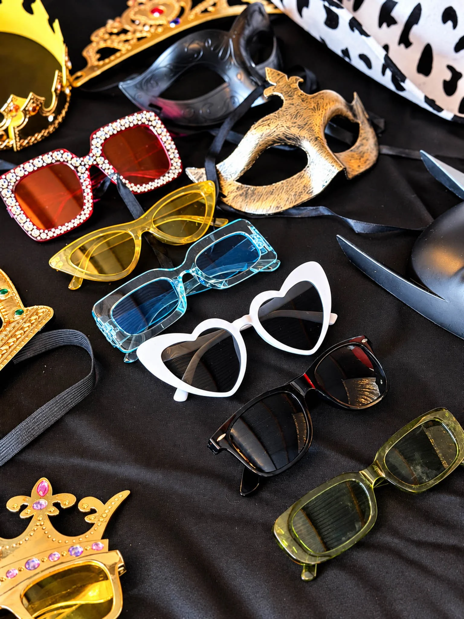 Selection of colorful photo booth glasses, masks, and jeweled crowns arranged on a black tablecloth