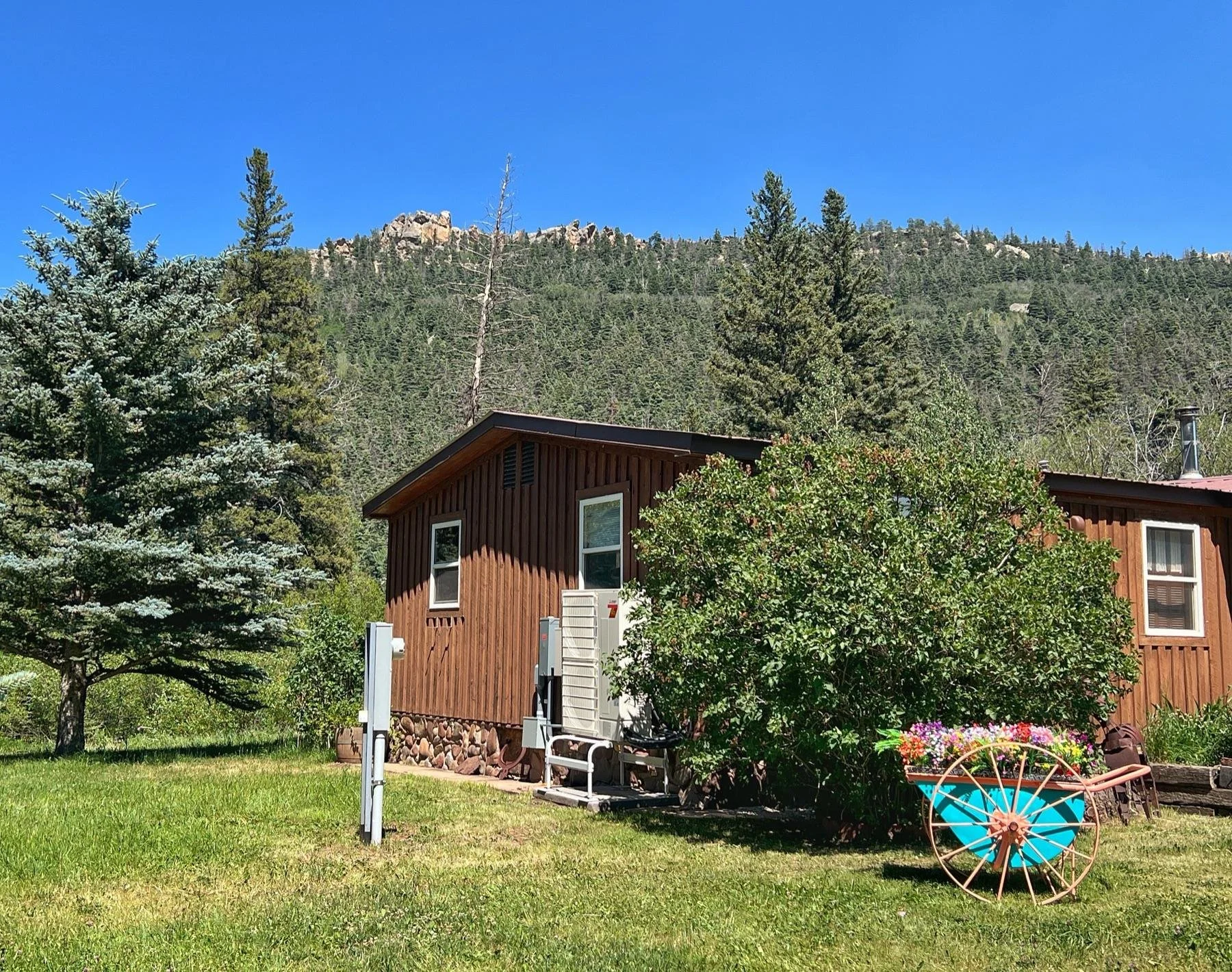 Brown wood cabin with a Mitsubishi outdoor unit, pine trees and a rocky ridge behind it