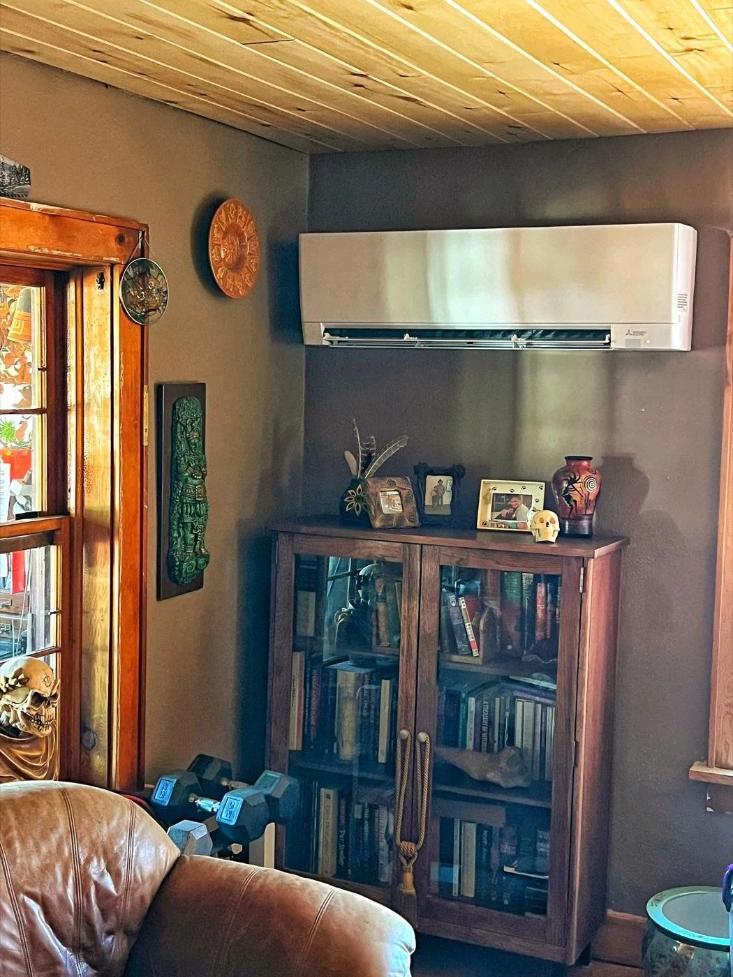 Wall-mounted Mitsubishi indoor unit above a bookcase in a living room with a wood ceiling
