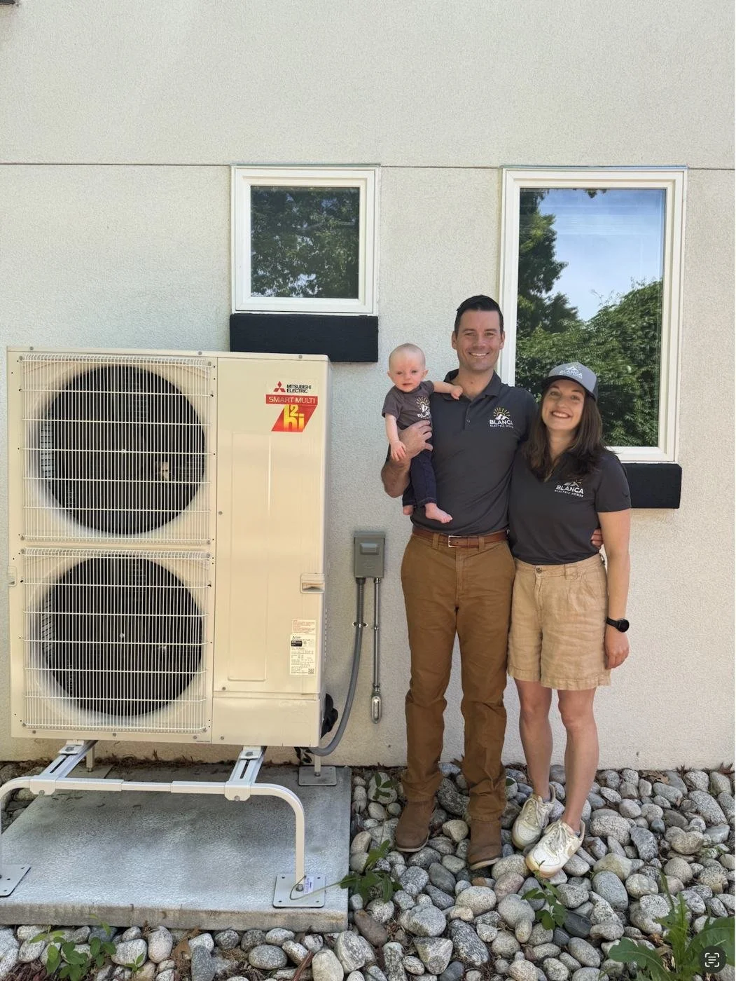 Blanca's owners, holding their baby, standing beside a Mitsubishi outdoor heat pump unit