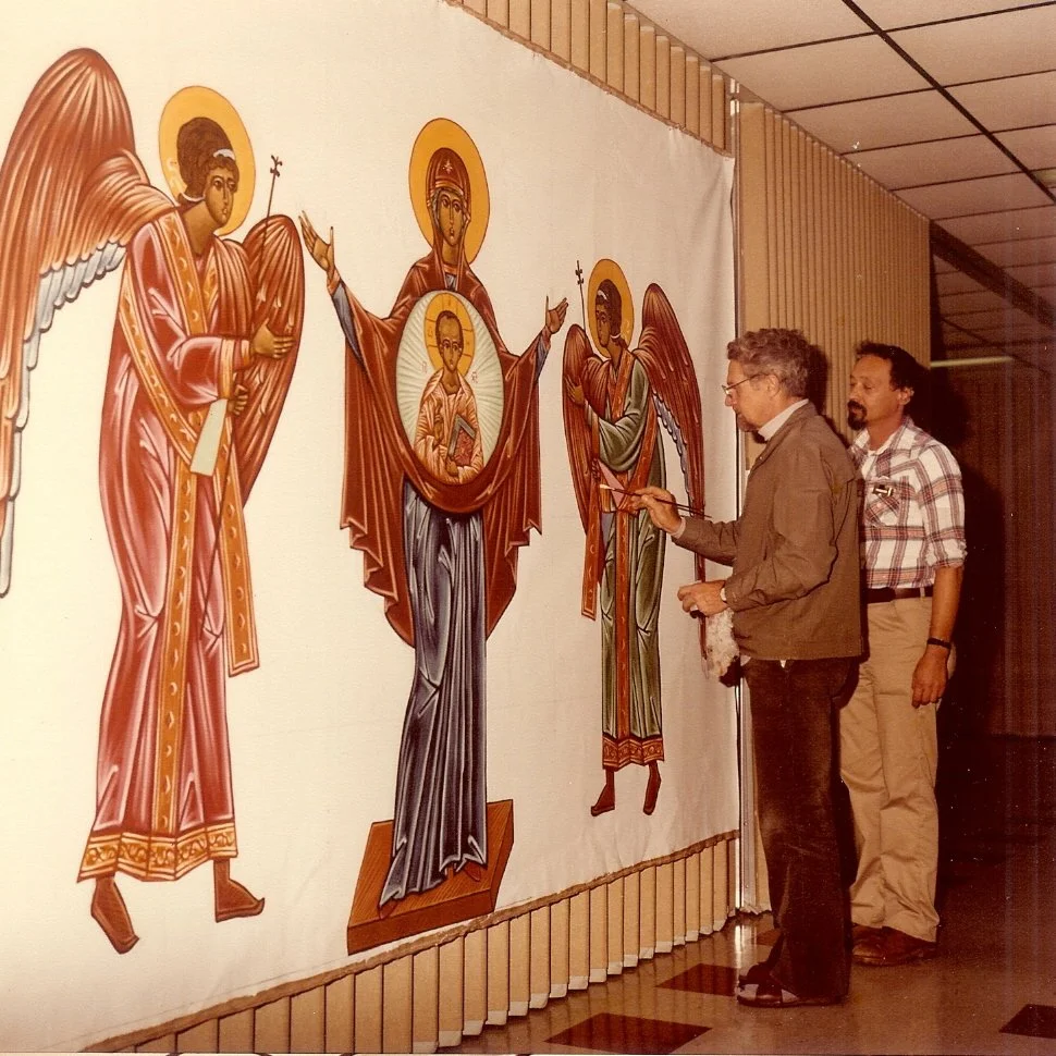 An iconographer painting an icon of the Theotokos and Christ Child flanked by angels, with another man looking on