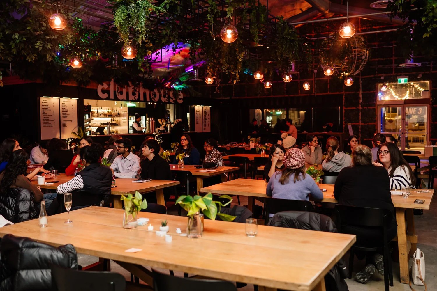 A full room of people talking at long tables under string lights