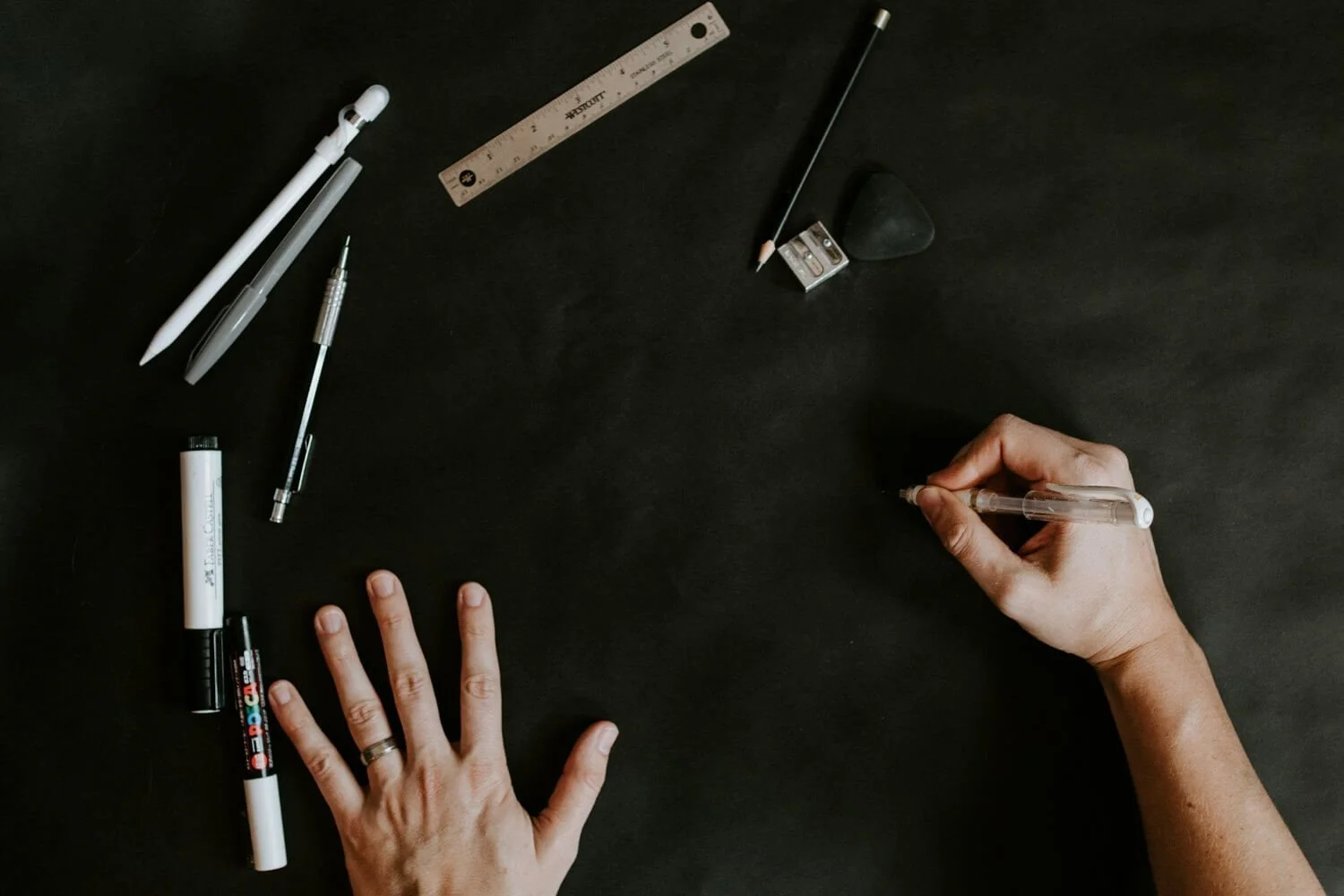 A hand sketching beside drafting tools on a dark surface