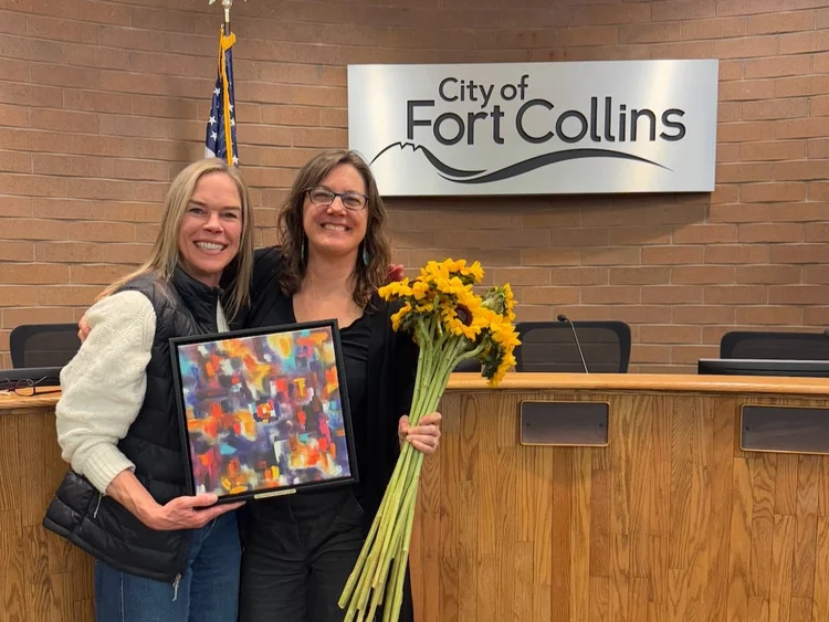 Photo of two women embracing holding a picture and flowers in front of a desk.