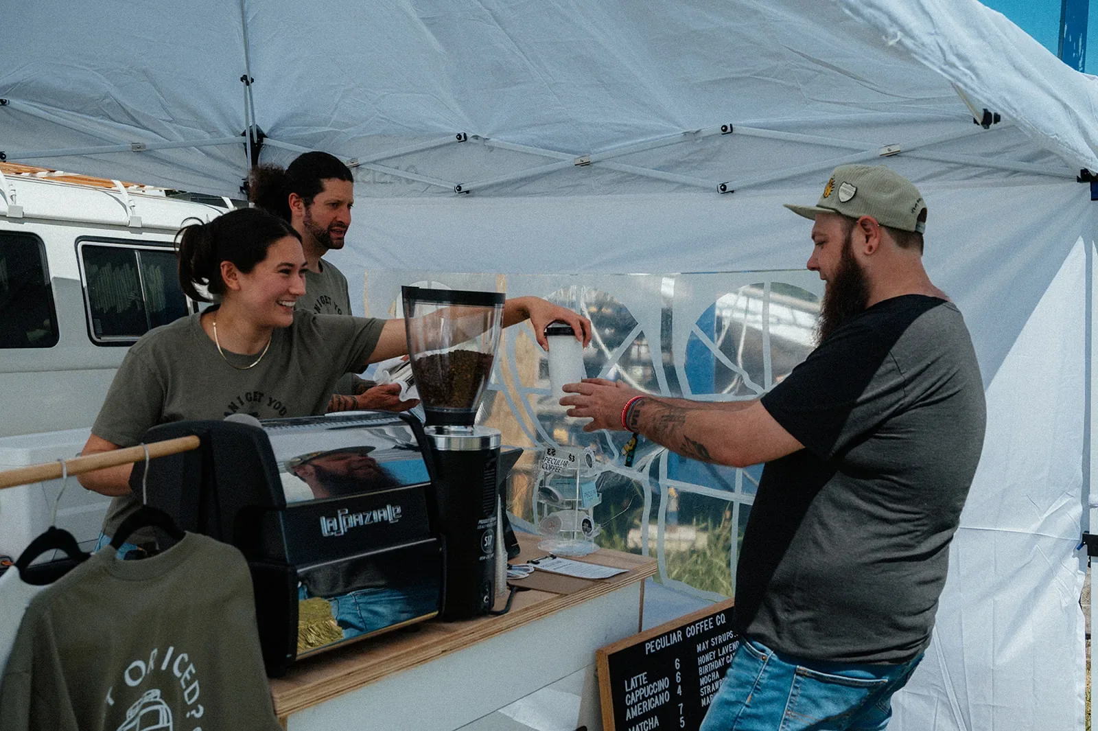 Smiling Peculiar Coffee baristas handing a fresh drink to a happy guest under the event tent