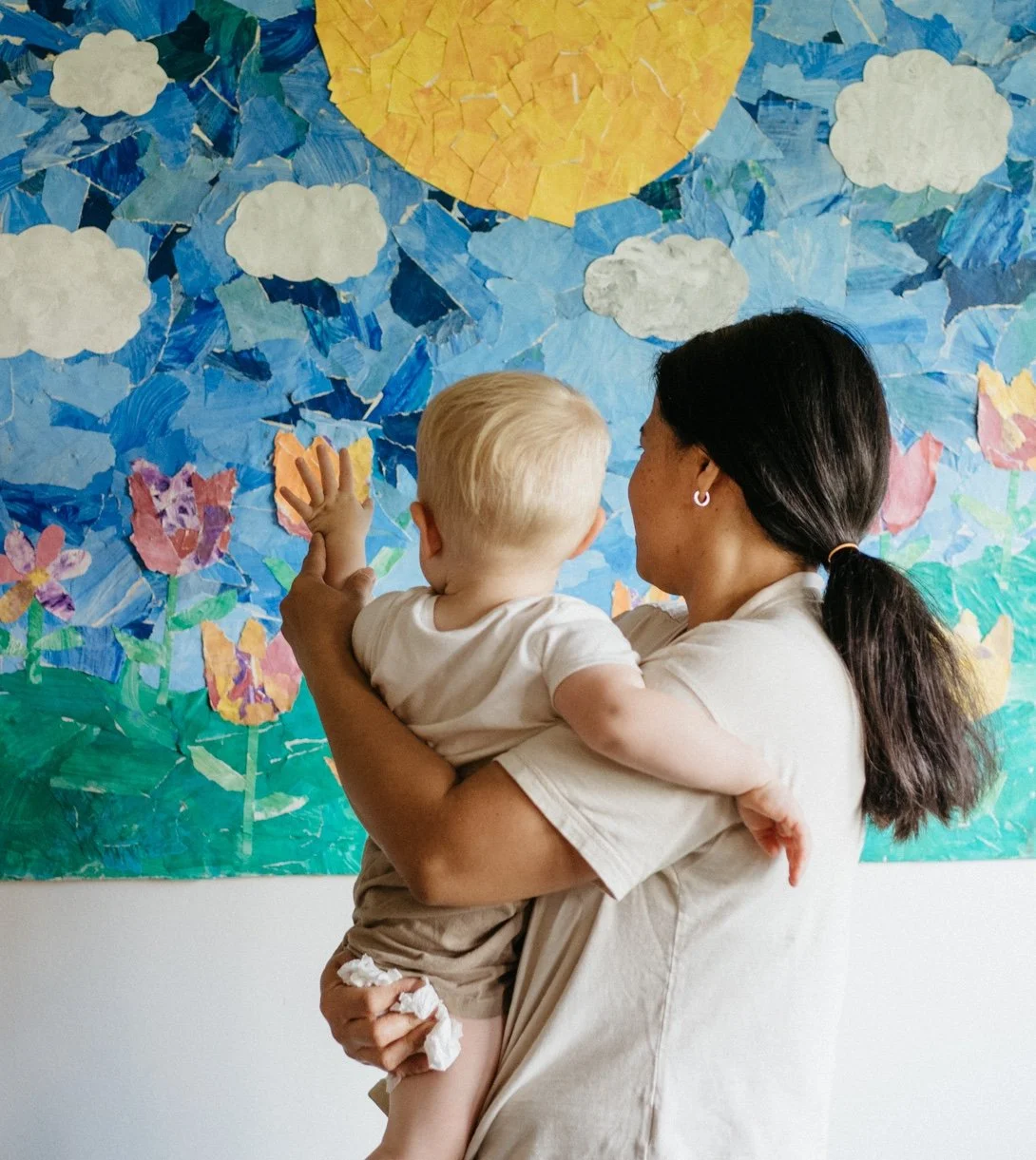 An educator holding a child as they look at a handmade sun-and-flowers mural