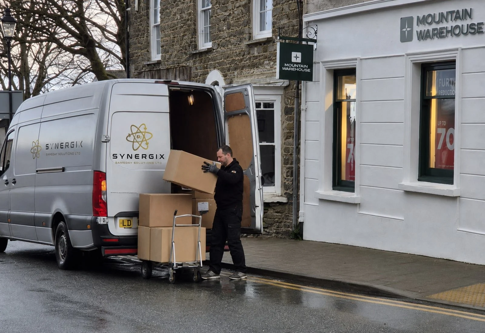 Delivery worker unloading cartons from a Synergix-branded van beside a retail store.