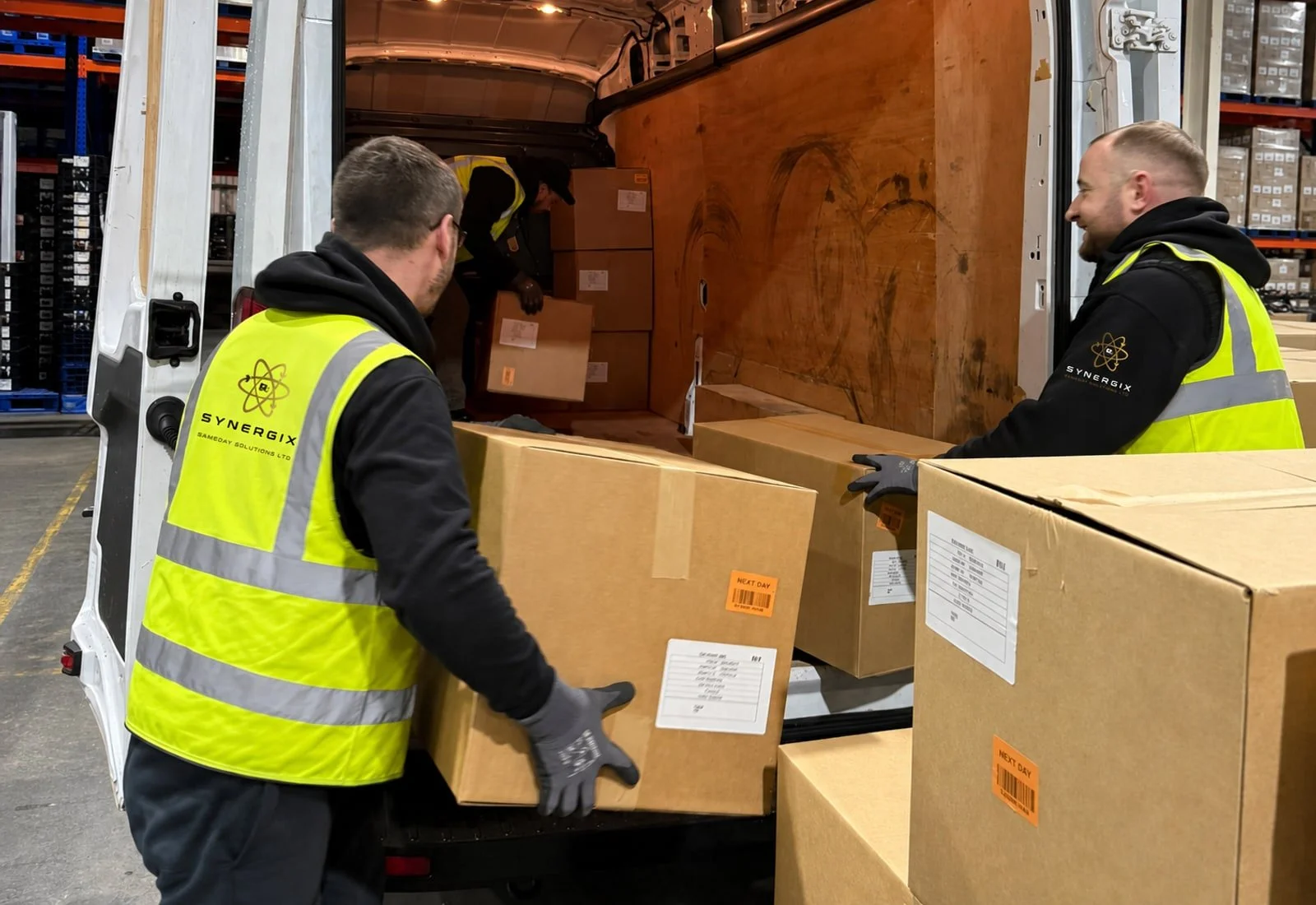 Two-person delivery team in Synergix-branded workwear handling cartons at the rear of a van.