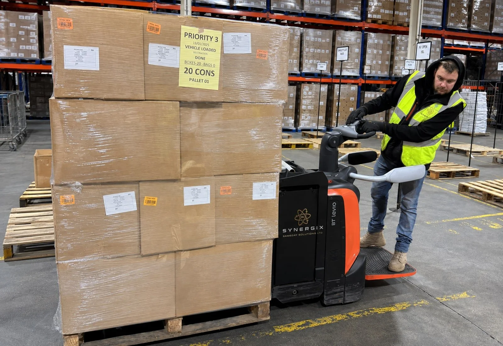 Warehouse worker in Synergix-branded workwear moving a wrapped pallet with an electric pallet truck.