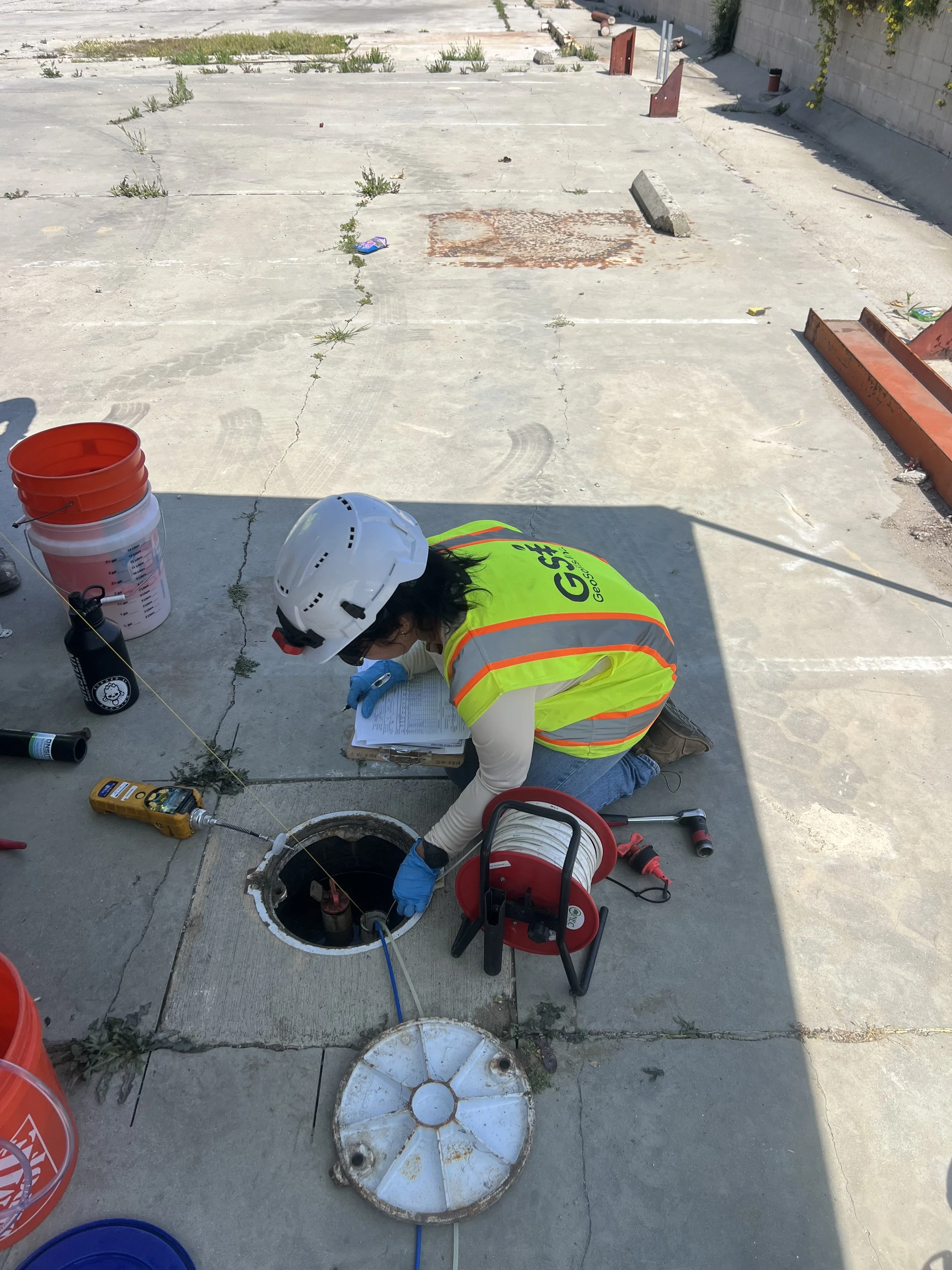 Environmental field technician in hard hat and safety vest sampling a groundwater monitoring well at an industrial site, recording field measurements