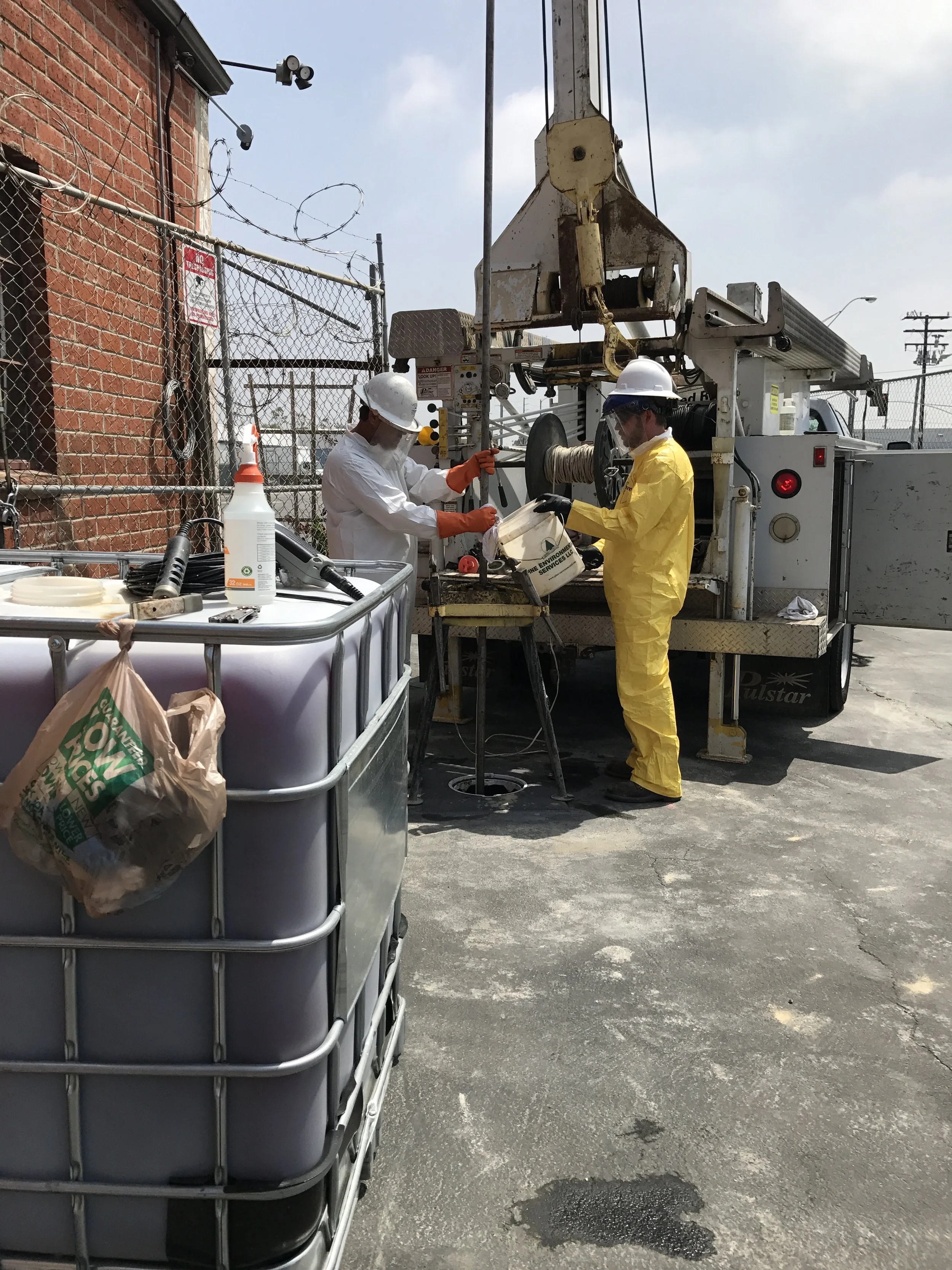 Environmental drilling crew in Tyvek suits and chemical-resistant PPE operating a direct push drill rig at a hazardous materials investigation site