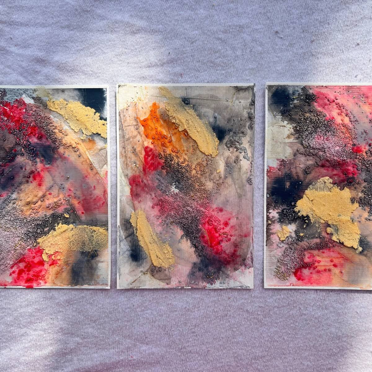 Three artworks from the “And The Fire Within” series on fabric – glowing red, gold and black tones in sunlight, inspired by inner rage and transformative power.