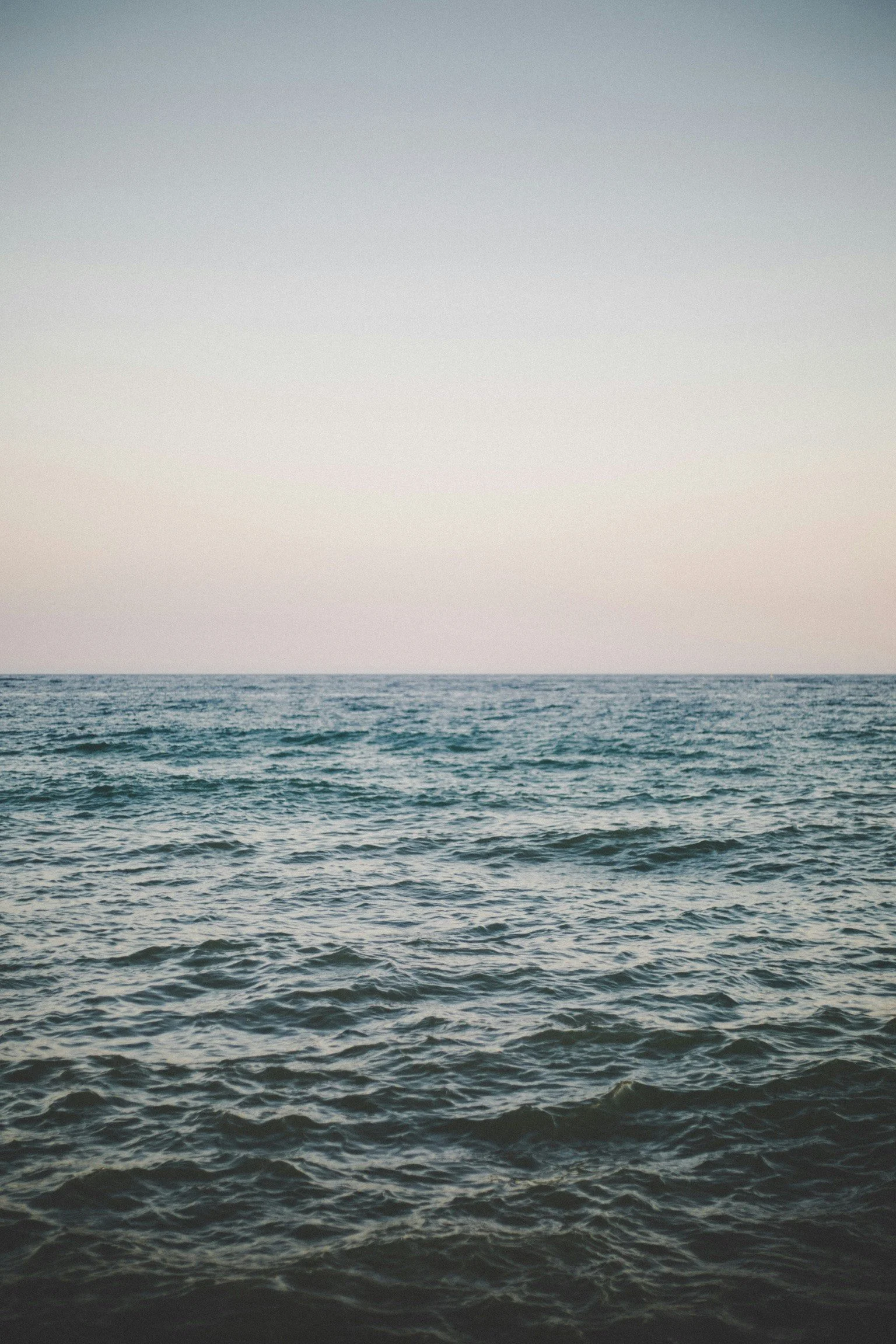 A calm ocean at dusk, meeting a pale sky at the horizon