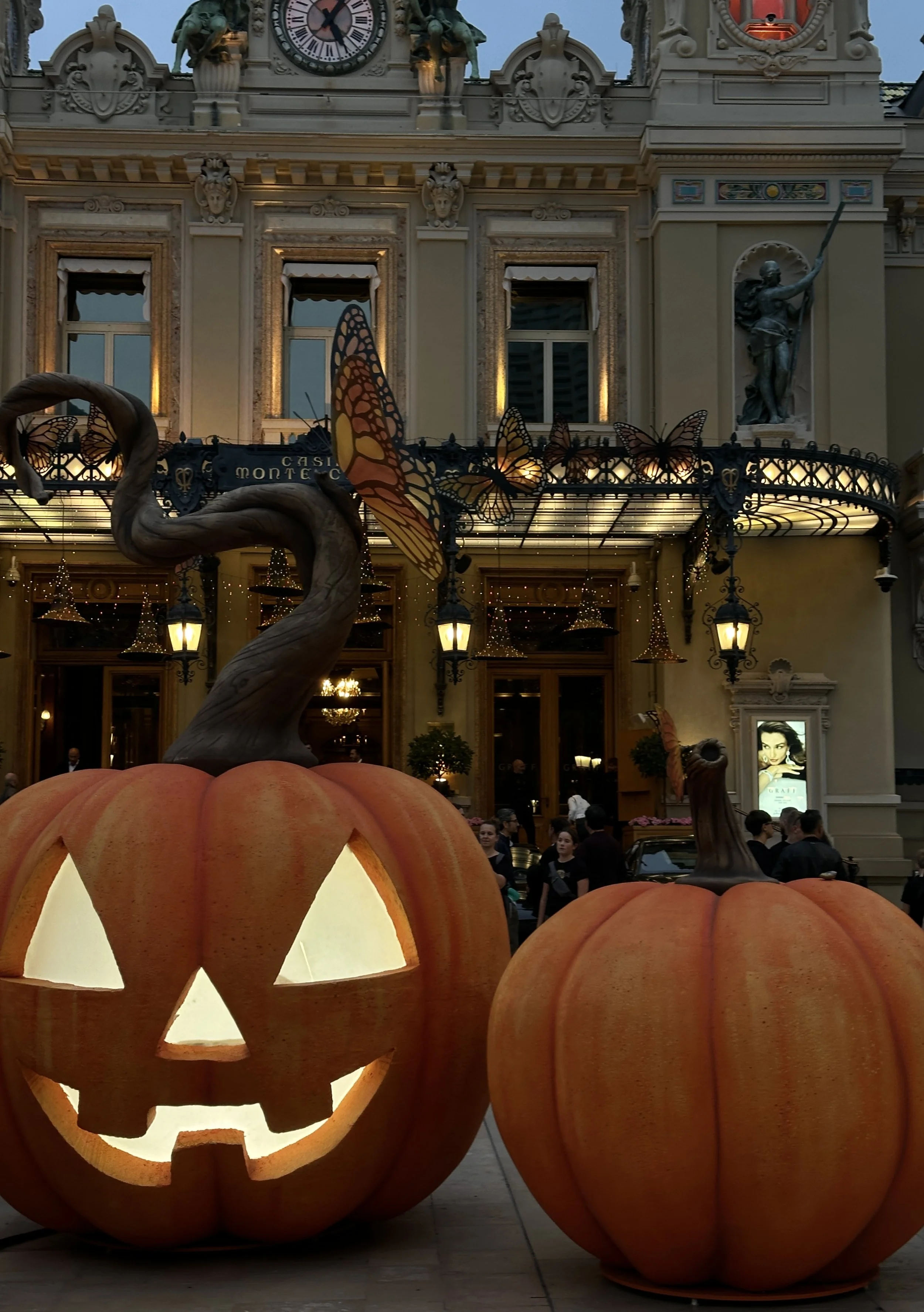 Two giant illuminated jack-o'-lantern pumpkins at the entrance of the Casino de Monte-Carlo at dusk, with witch-hat lanterns suspended from the veranda above