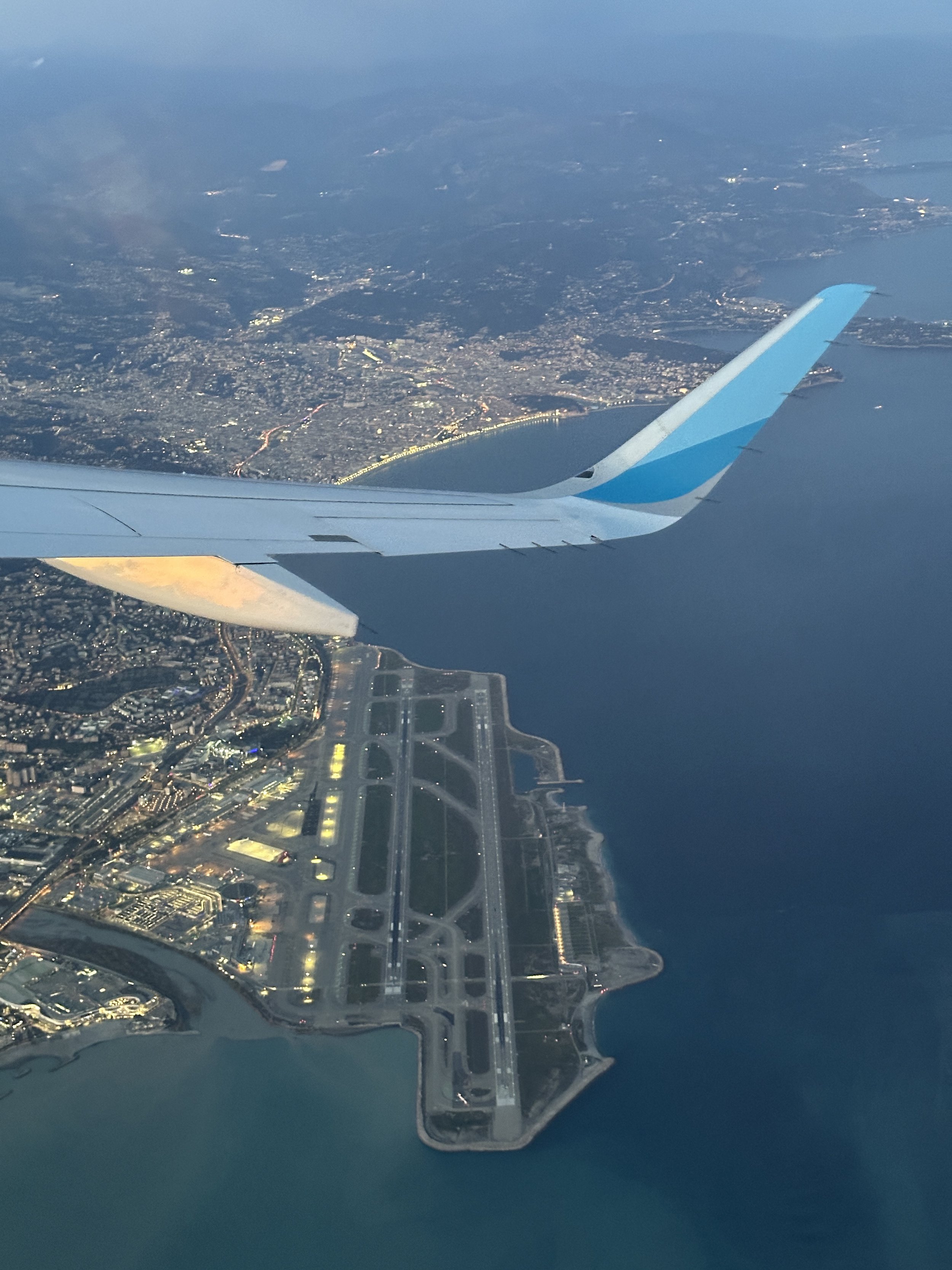 Nice Côte d'Azur Airport seen from above at dusk — the airport's twin parallel runways extending into the Mediterranean, the city of Nice lit up along the Promenade des Anglais, aircraft wing visible