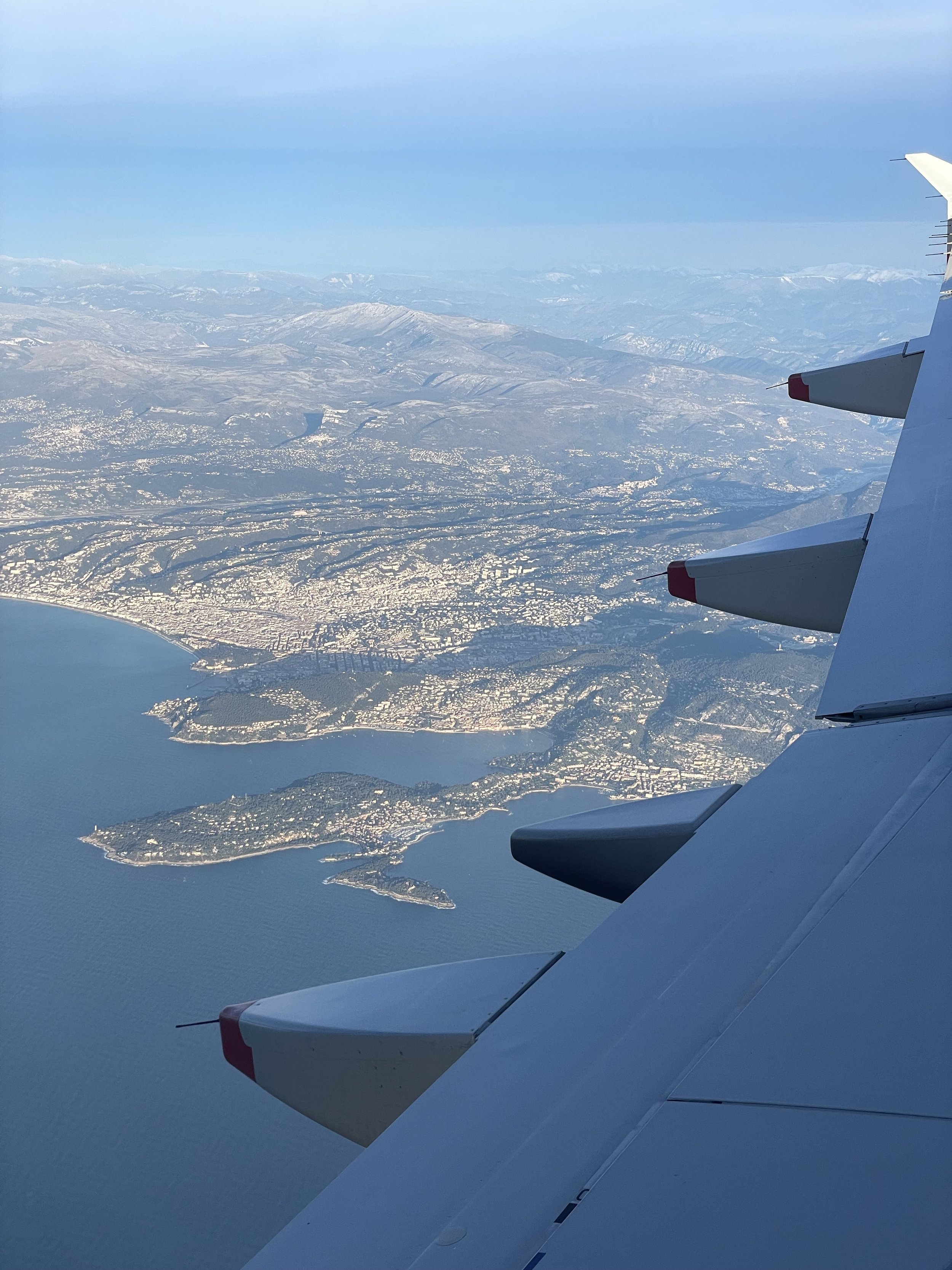 View from an aircraft window on approach to Nice — Cap d'Antibes peninsula and the Riviera coastline below with snow-capped Alps rising in the distance behind