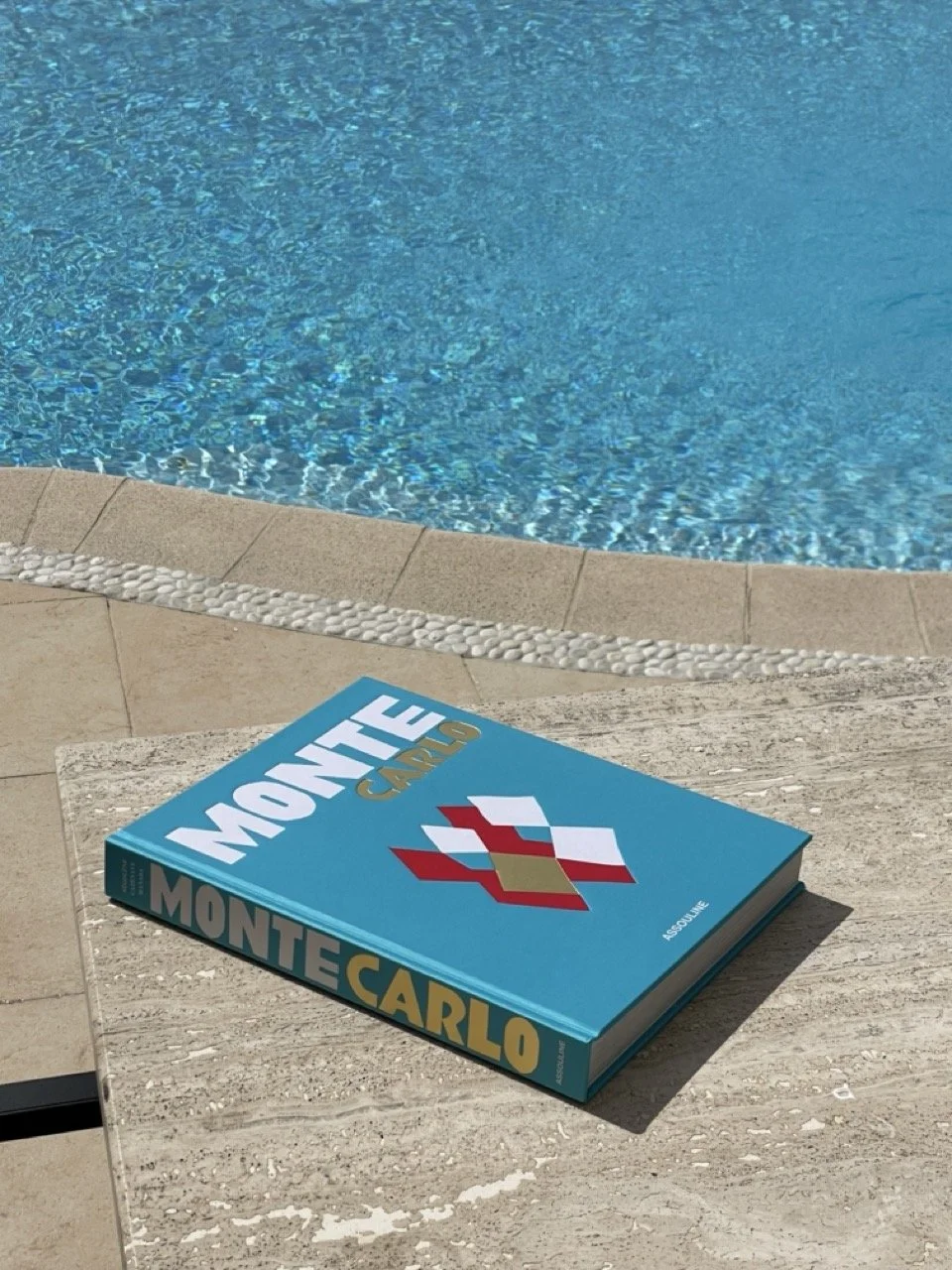 An Assouline Monte Carlo coffee table book with its distinctive turquoise cover resting on the travertine edge of Villa Monaco's pool