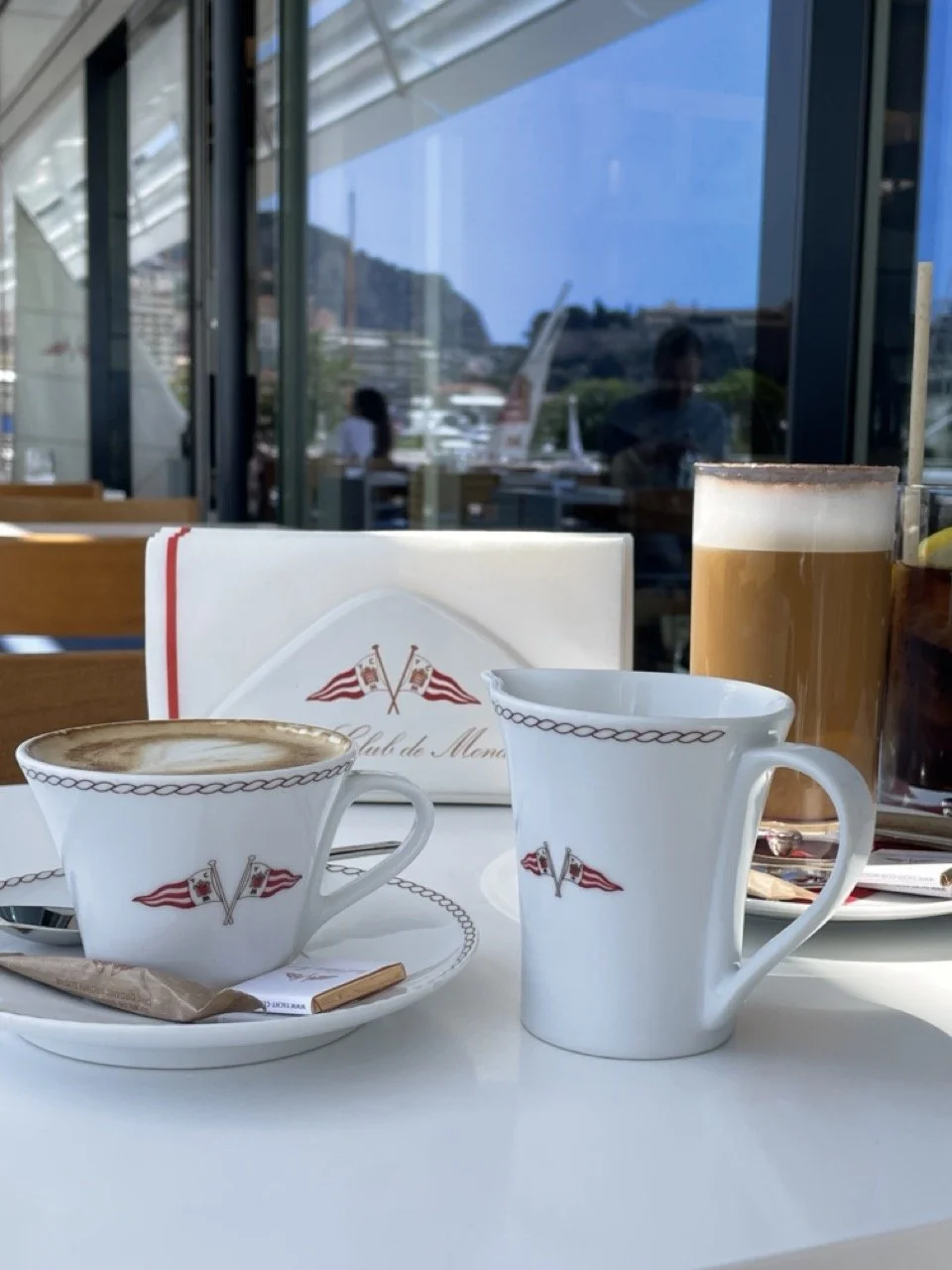 Coffee service at the Yacht Club de Monaco with the club's red and white burgee on the porcelain, and Port Hercule and the Rocher visible through the window