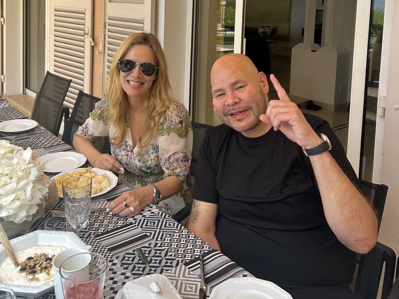 Villa Monaco owner Jessica Stockmann and rapper Fat Joe seated together at an outdoor breakfast table at the villa, with white hydrangeas and a black-and-white geometric table runner