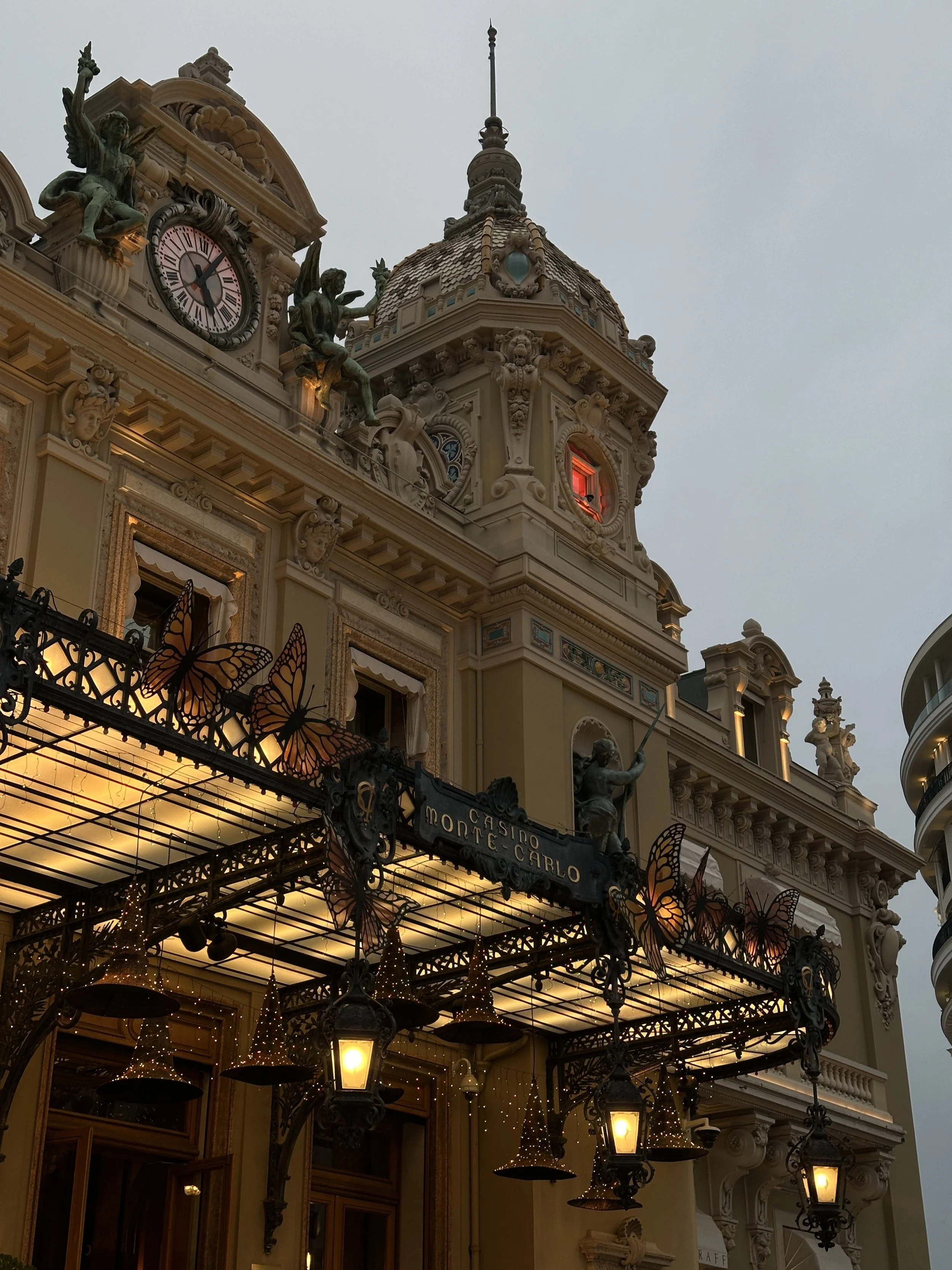 The Belle Époque façade of the Casino de Monte-Carlo at dusk, dressed for Halloween with giant monarch butterfly decorations on the wrought-iron veranda and witch-hat lanterns suspended beneath