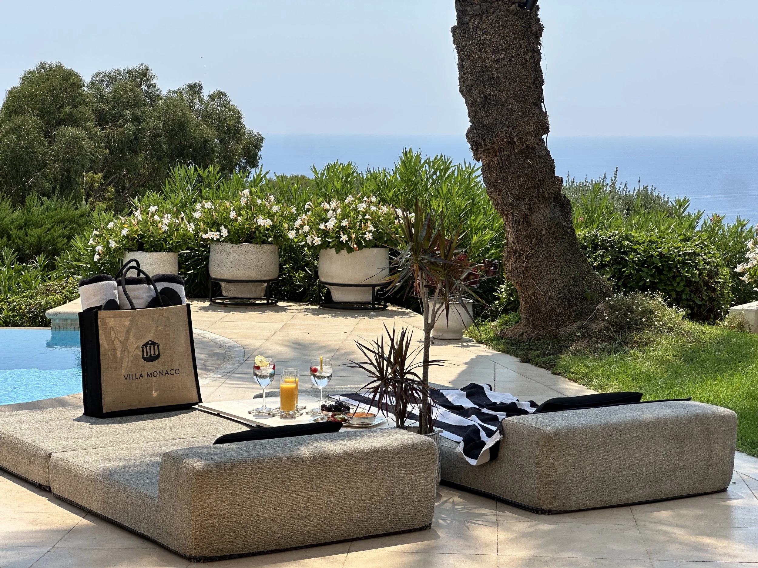 Villa Monaco's poolside sun-loungers with a branded jute bag and a small selection of cocktails and fruit set out beside the infinity pool