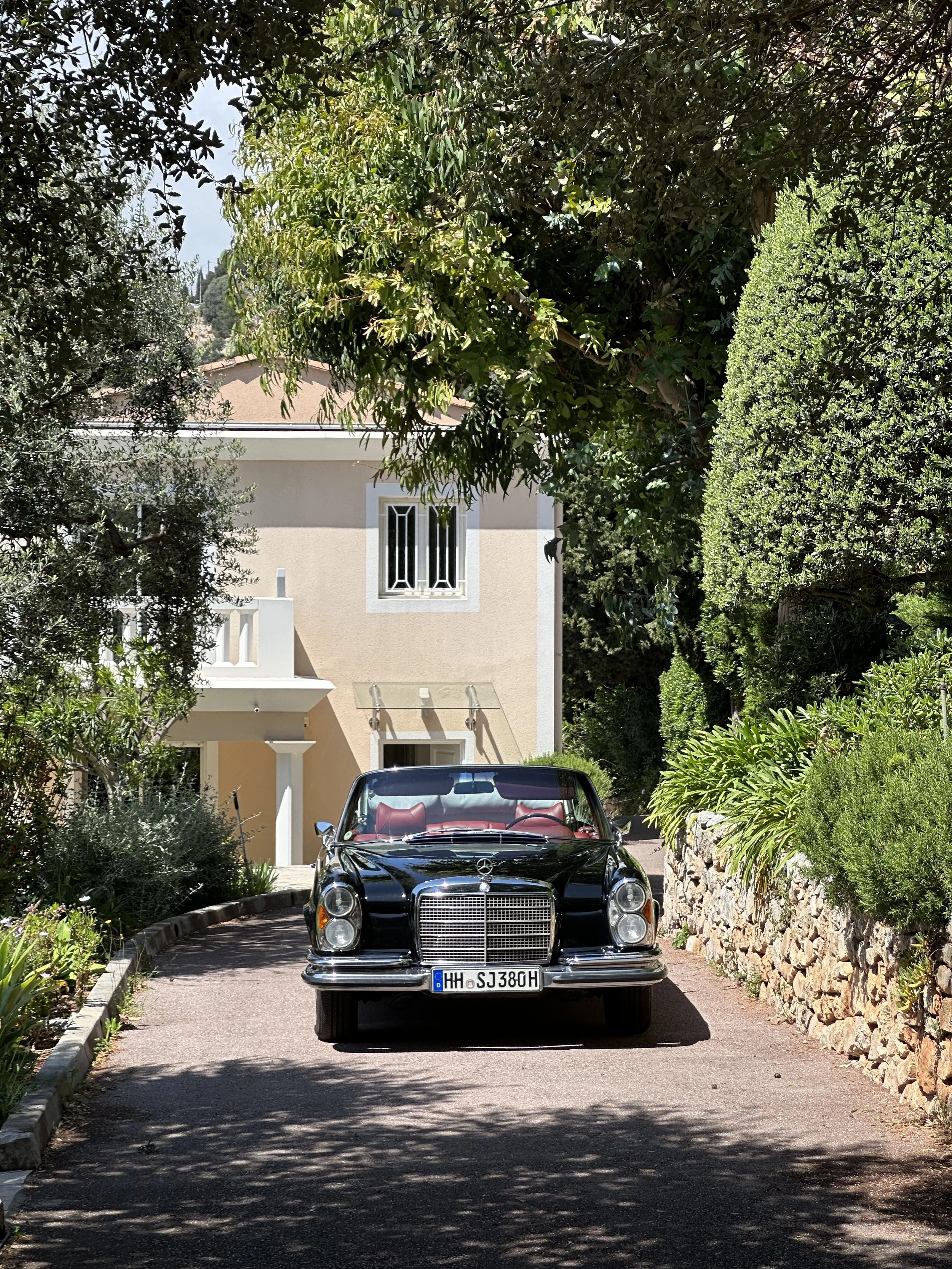 A dark classic Mercedes-Benz W111 Cabriolet with red leather interior and Hamburg historic-vehicle registration parked at the Villa Monaco entrance, framed by olive trees, cypress and a dry-stone wall