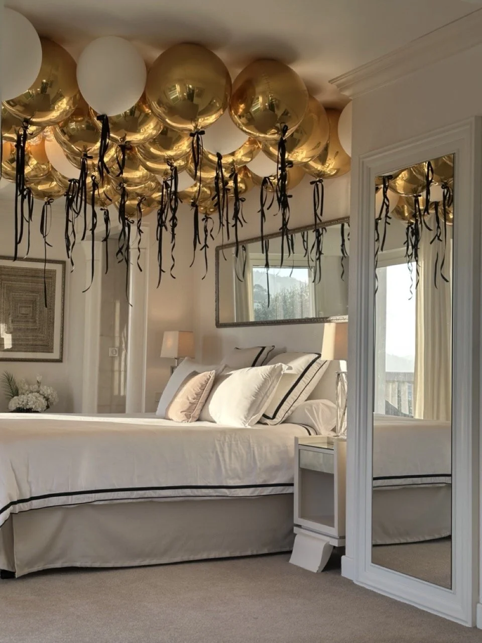 A Villa Monaco master bedroom with the king bed dressed in cream and white linens topped with a canopy of gold and cream balloons trailing black satin ribbons — a surprise arrival for the birthday guest of honour