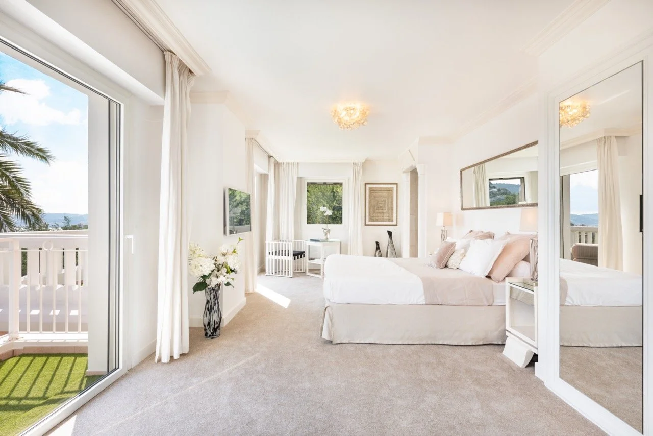 A master bedroom at Villa Monaco with a low upholstered platform bed in cream, matching bedside lamps, and floor-to-ceiling windows opening to the Mediterranean light
