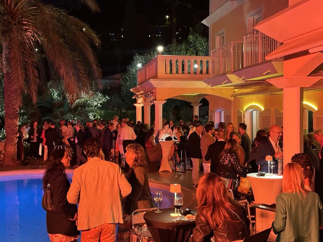 An evening cocktail reception at Villa Monaco with guests gathered around cocktail tables on the illuminated terrace, the villa's arched façade and palm trees behind