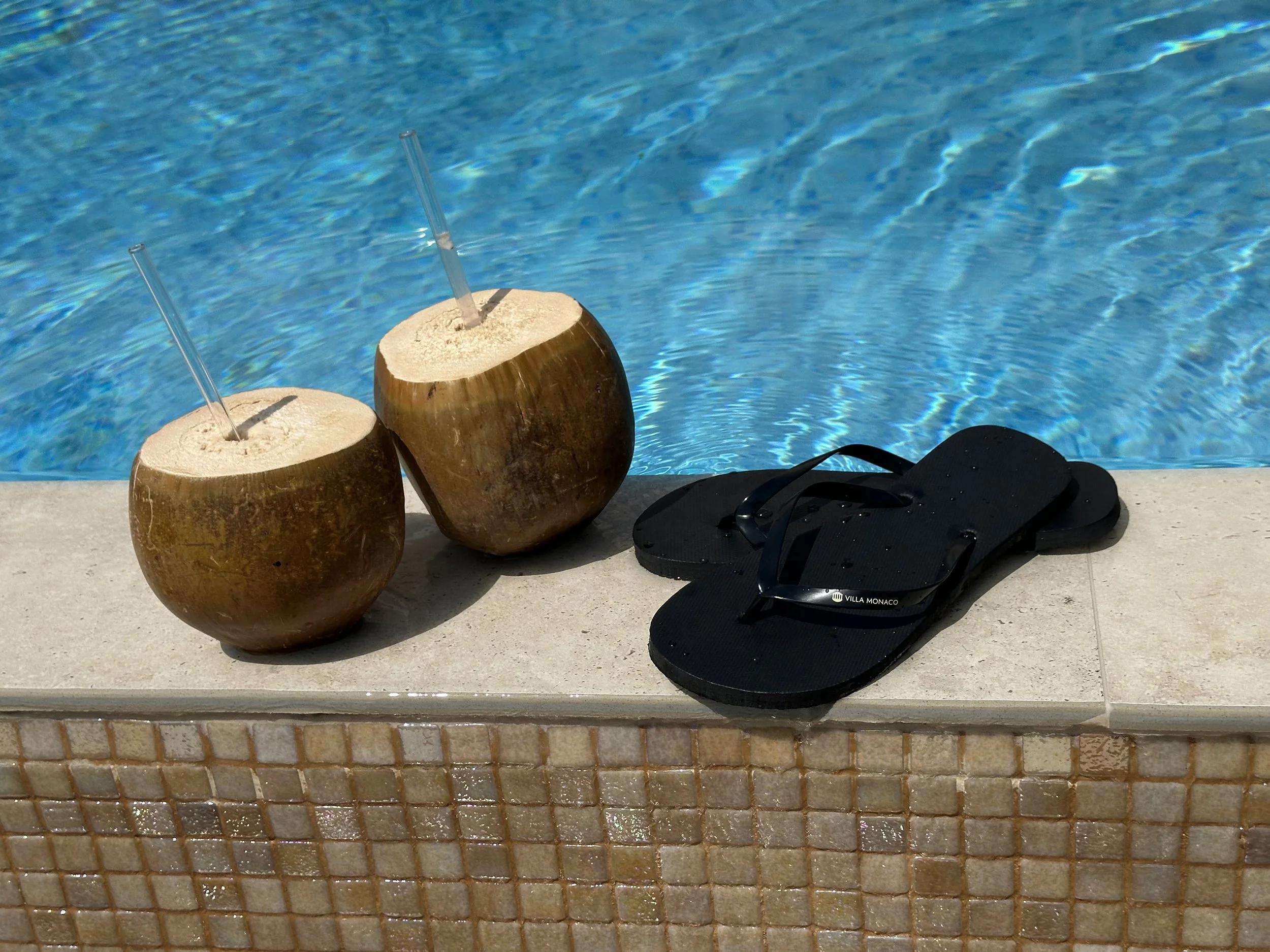 Two fresh coconuts with straws and a pair of Villa Monaco branded black flip-flops beside the villa's private pool