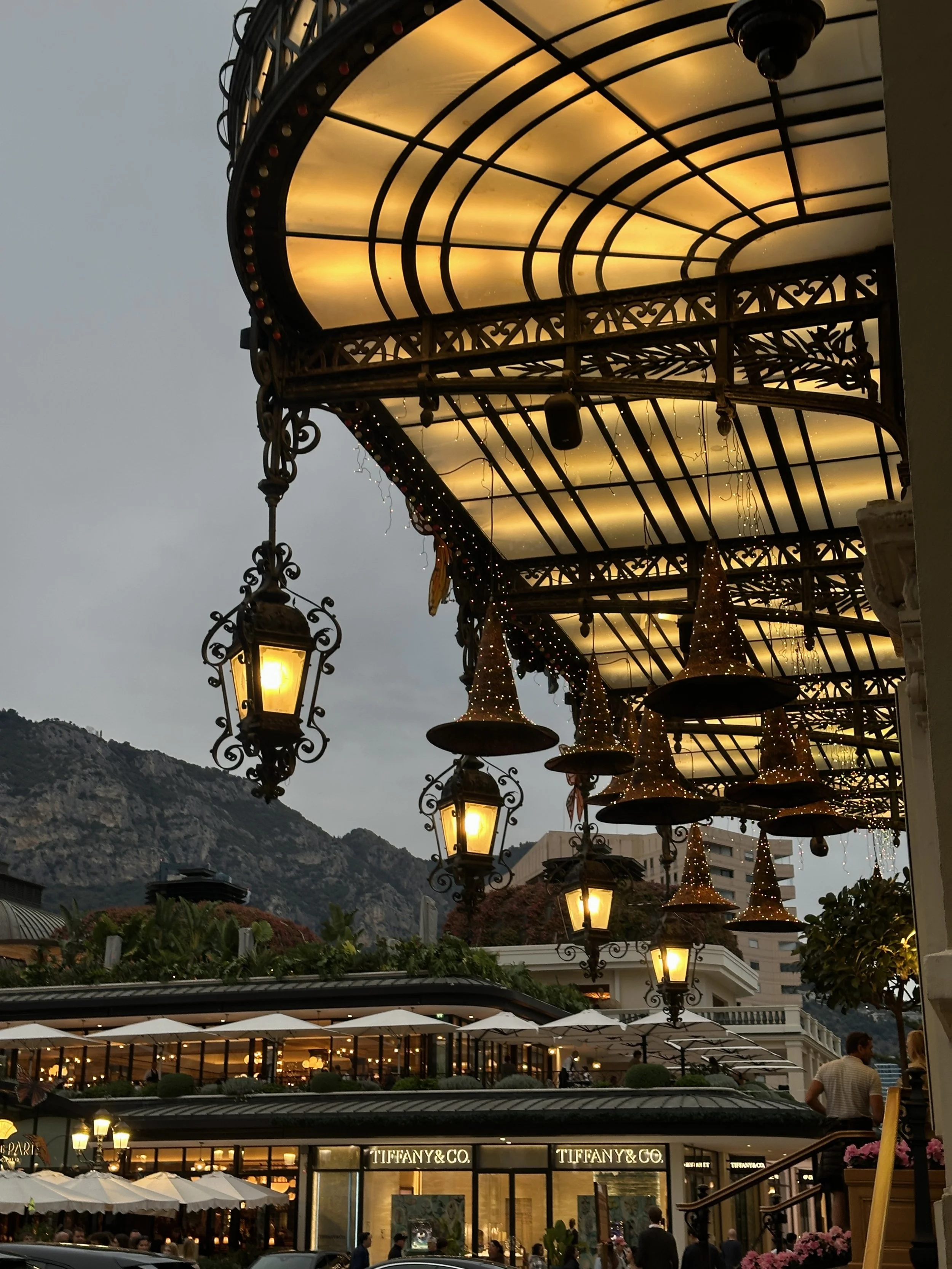 Illuminated witch-hat lanterns suspended beneath the ornate wrought-iron veranda of the Casino de Monte-Carlo, with Tiffany & Co. and Hôtel de Paris café terrace visible across the square at blue-hour dusk