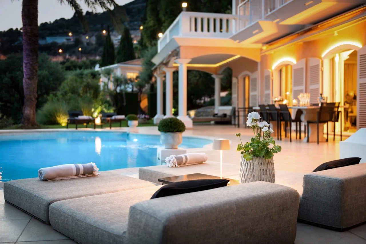Villa Monaco poolside at dusk with warm amber lighting on the arched façade, illuminated pool, sun-loungers, and a set dining table under the terrace shutters