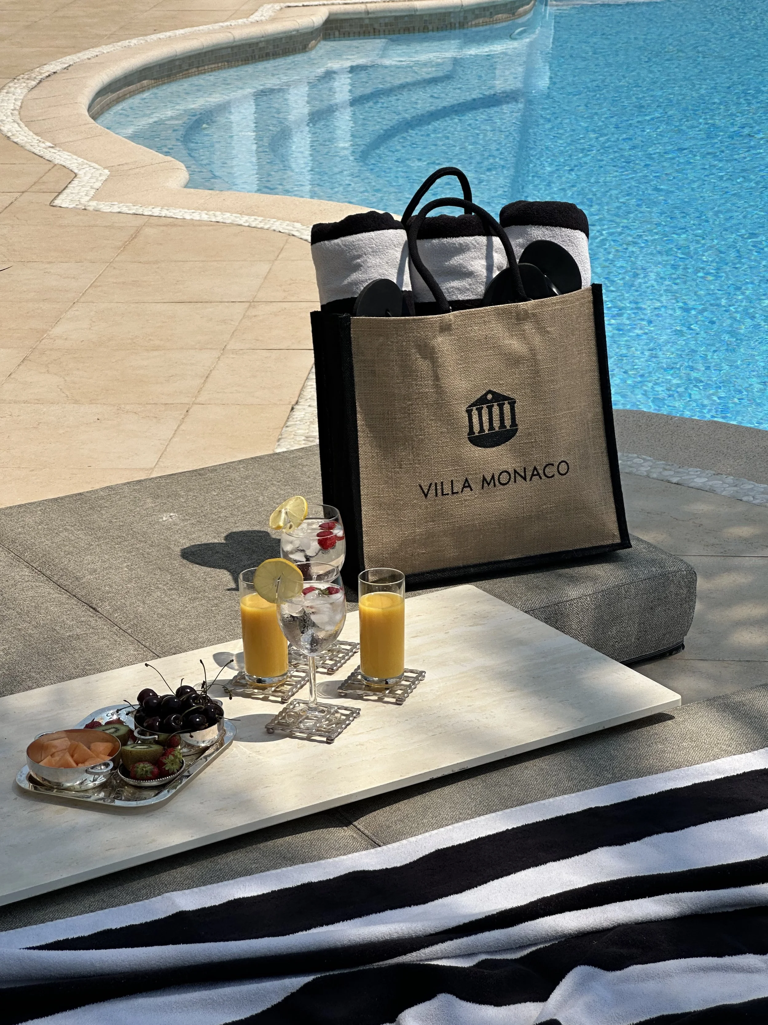 Villa Monaco's poolside styling — a branded jute bag with rolled black-and-white striped beach towels, cocktails and mocktails on a marble tray beside the infinity pool