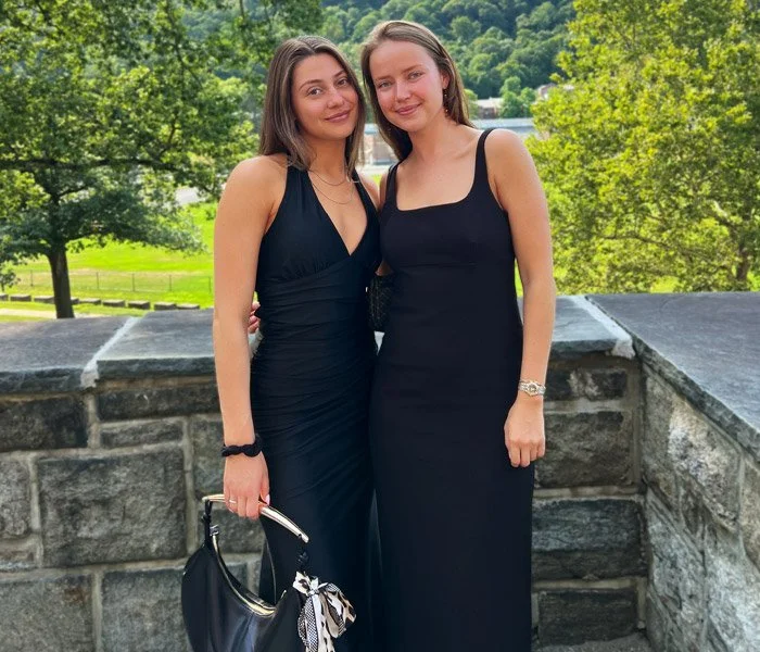 two tanned women wearing black dresses pictured outside by a stone wall
