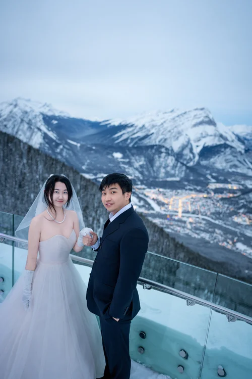 Couple in wedding attire at a glass railing with snow-covered mountains behind them